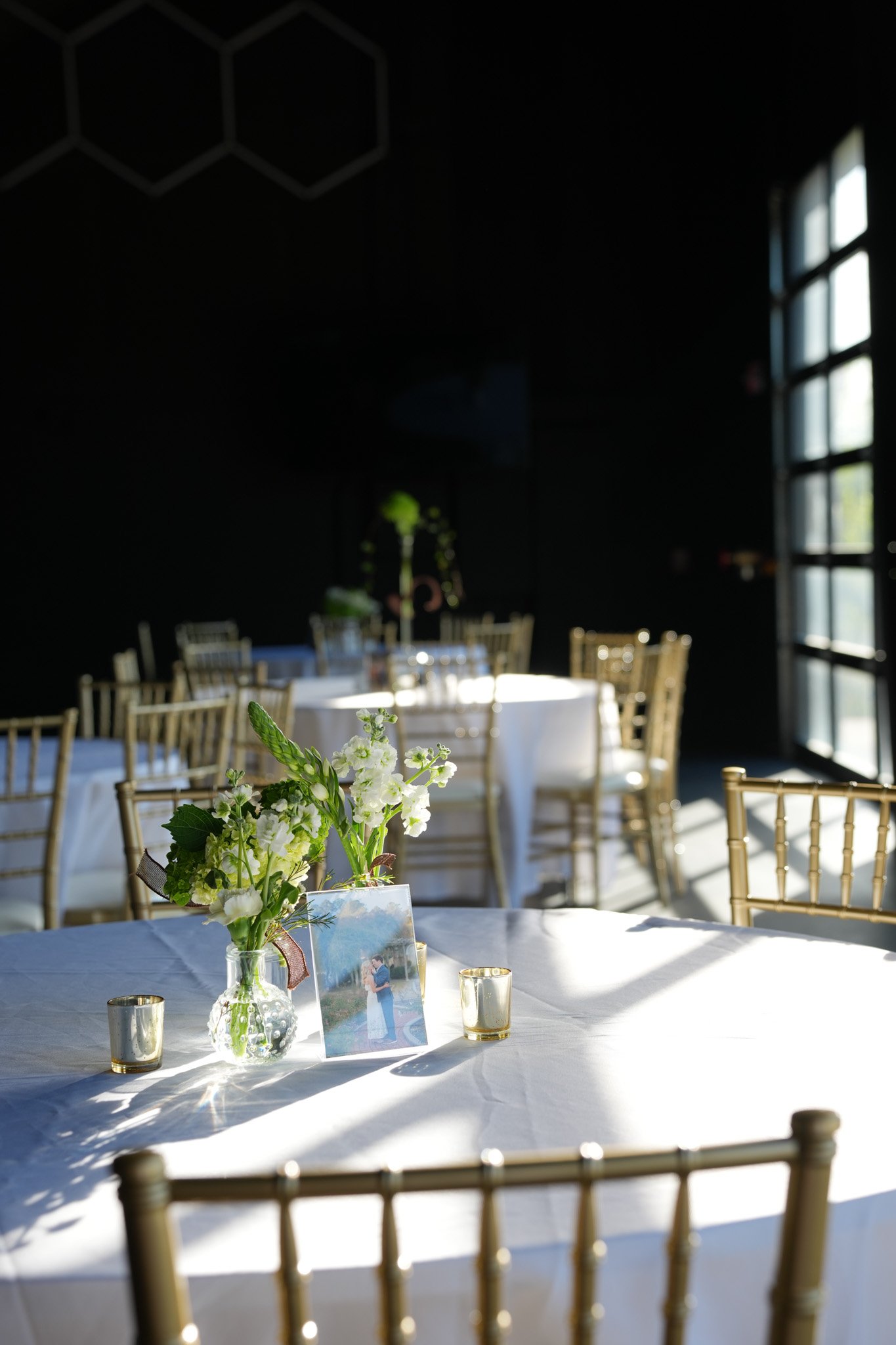 A table decorated with a floral centerpiece, framed photo, and gold-colored cups, set for an event in a room with large windows and natural sunlight.