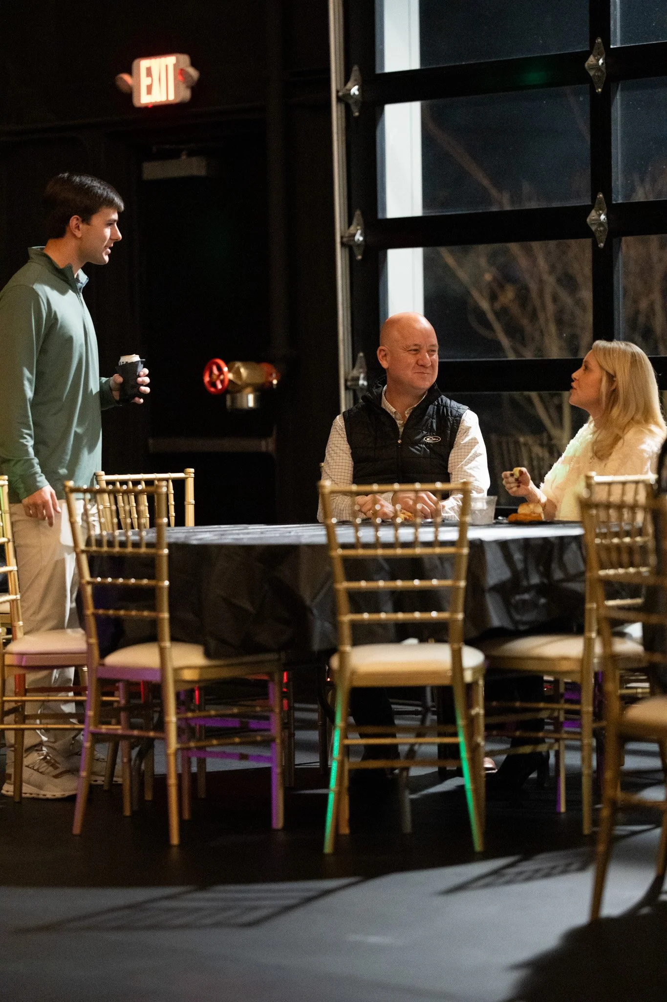 Three people in a dimly lit dining area, two women sitting at a table and a man standing with a coffee cup, engaging in conversation.