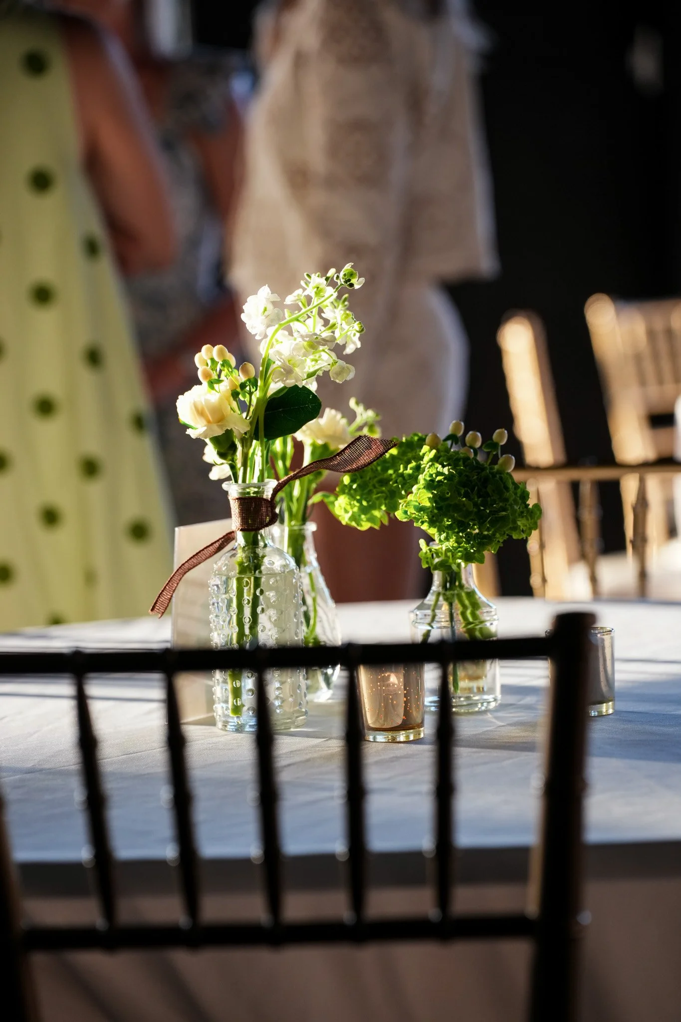 Close-up of white and green flowers in glass bottles on a table, with a blurred background and black chair back in the foreground.