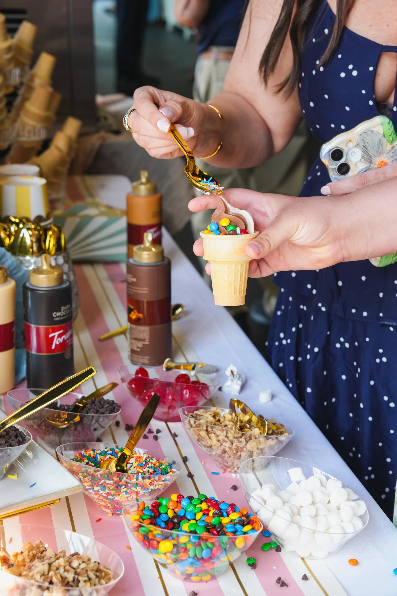 Close-up of a woman in a navy blue dress with white polka dots scooping colorful sprinkles into an ice cream cone at a dessert table with various toppings and containers.