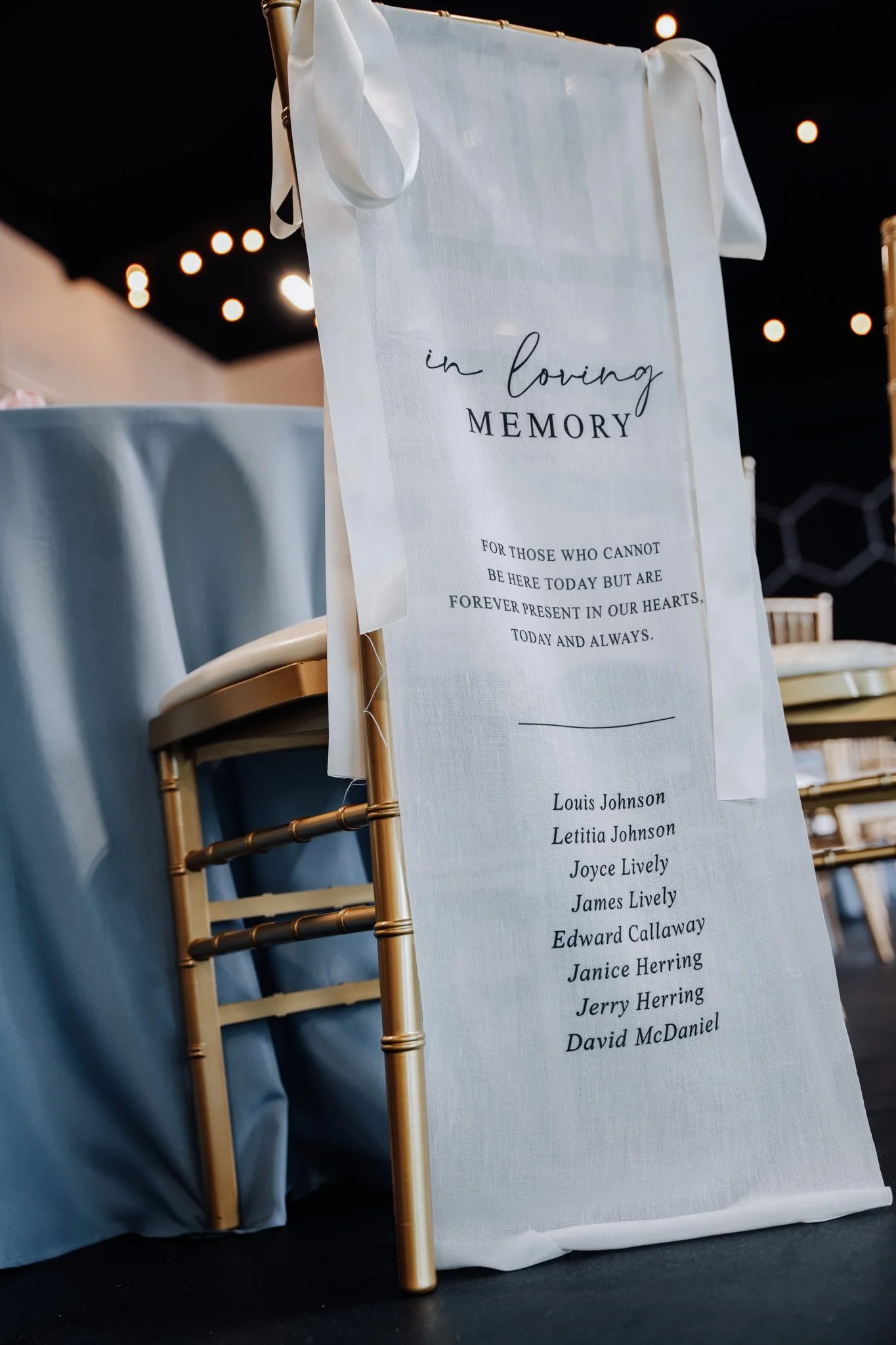 A memorial sign with a white fabric background and black text in a decorated venue. The sign reads 'in loving MEMORY' and includes a message for those who cannot be present. It lists several names at the bottom.