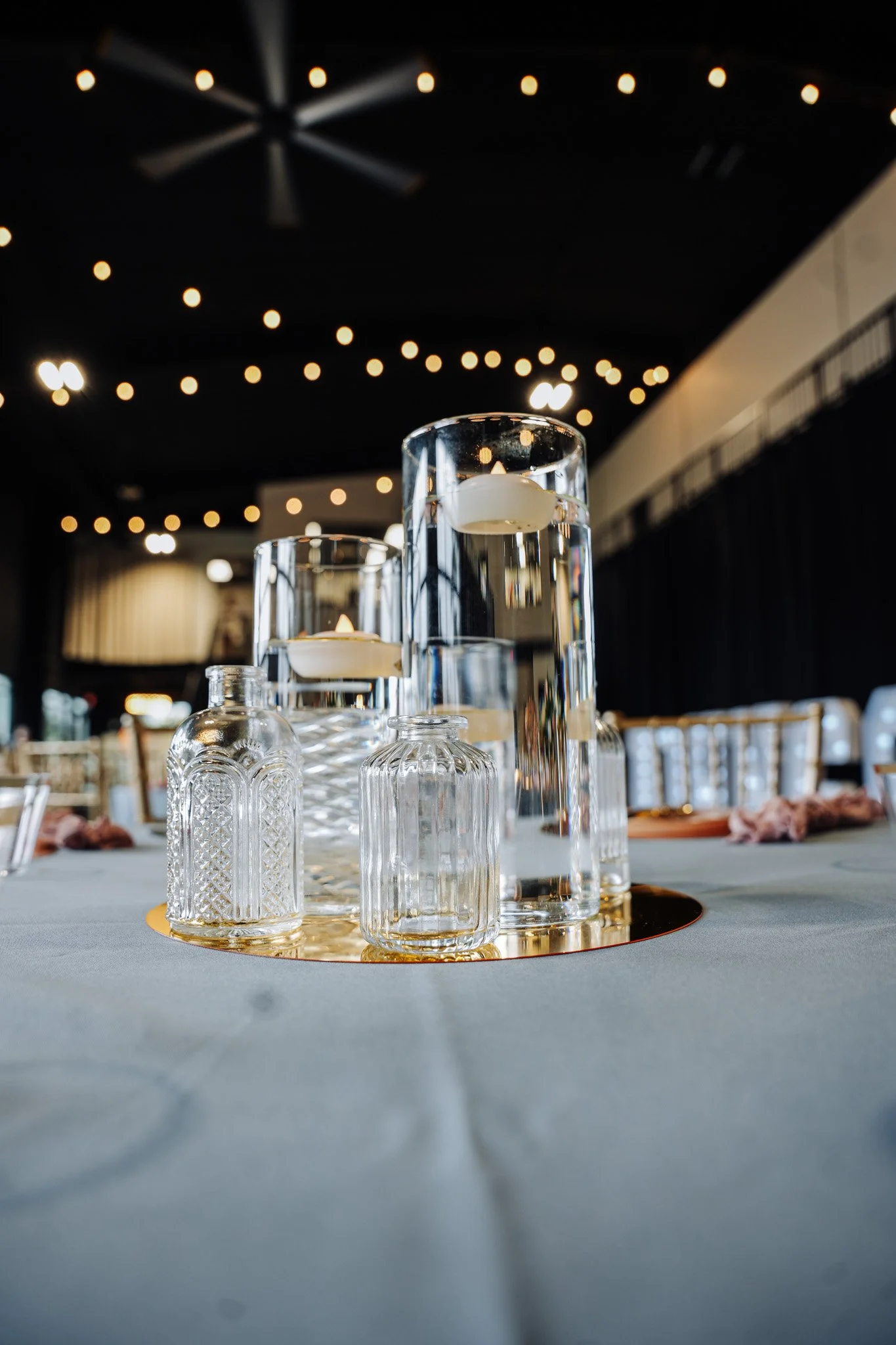 Glass candle holders with lit candles on a circular gold tray, set on a white tablecloth under hanging string lights in a dimly lit event hall.