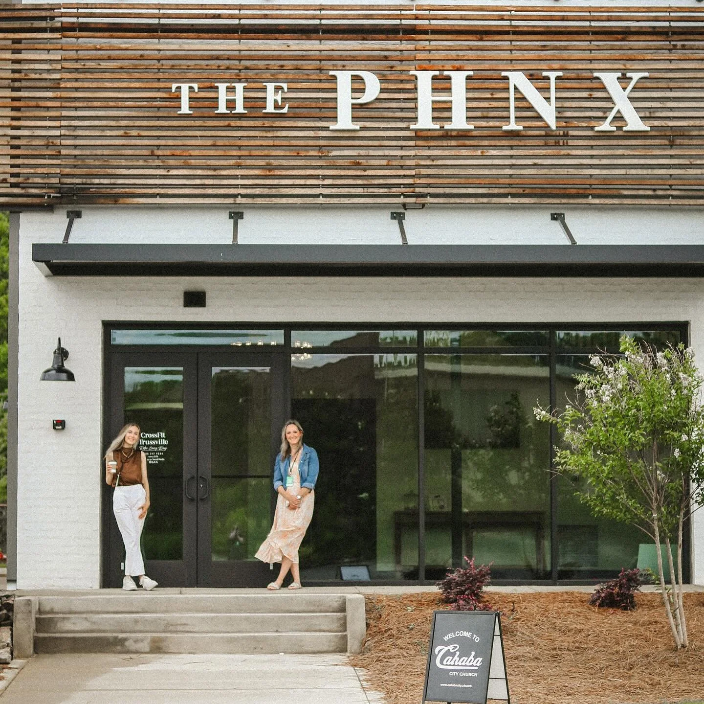 The exterior of a modern building with a sign reading 'The Pinh' and a smaller chalkboard sign that says 'Welcome to Cahaba City Church.' Two women are standing at the entrance, one holding a coffee cup.