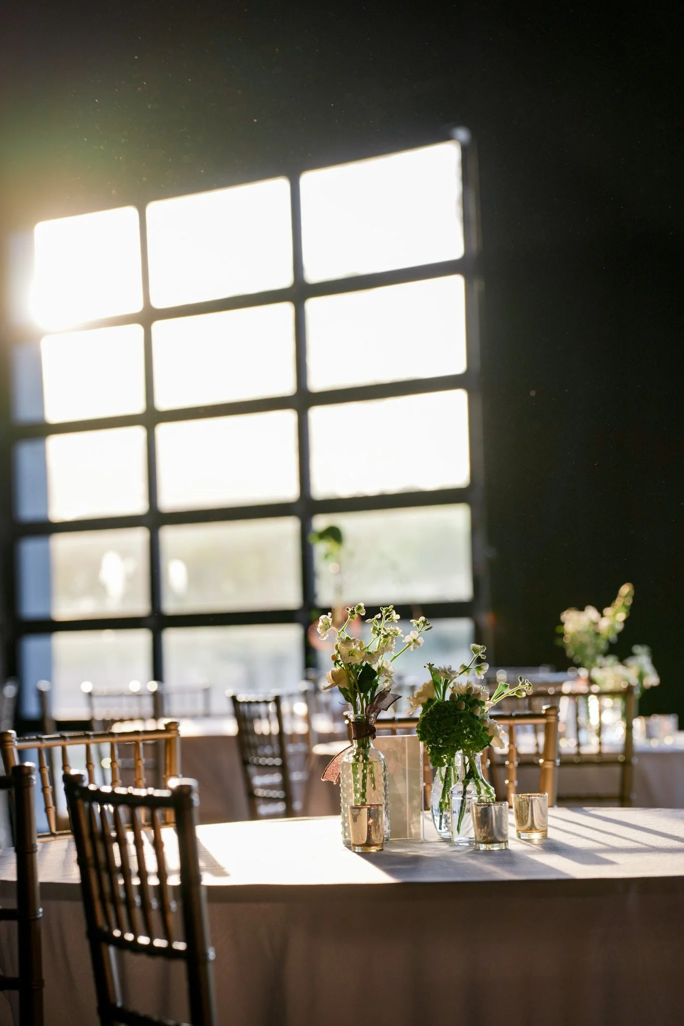 Sunlight streams through large window with grid panes into a dimly lit room decorated with flowers in vases on a table and surrounded by chairs