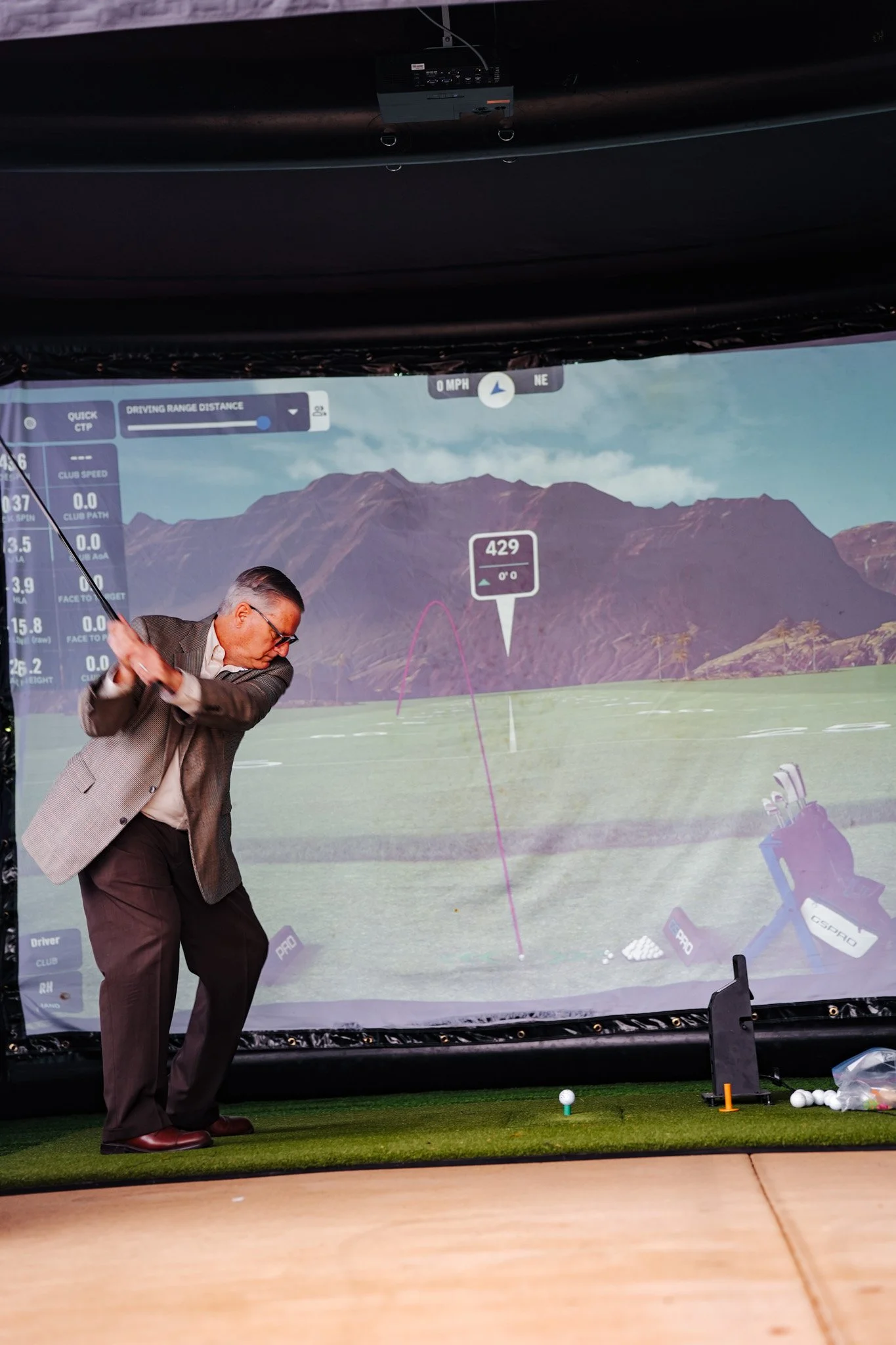 An elderly man in a gray plaid blazer and dark trousers practicing golf indoors on a simulator. He is holding a golf club following through after a swing, aiming at a virtual golf course projected on a large screen behind him. The screen displays mou