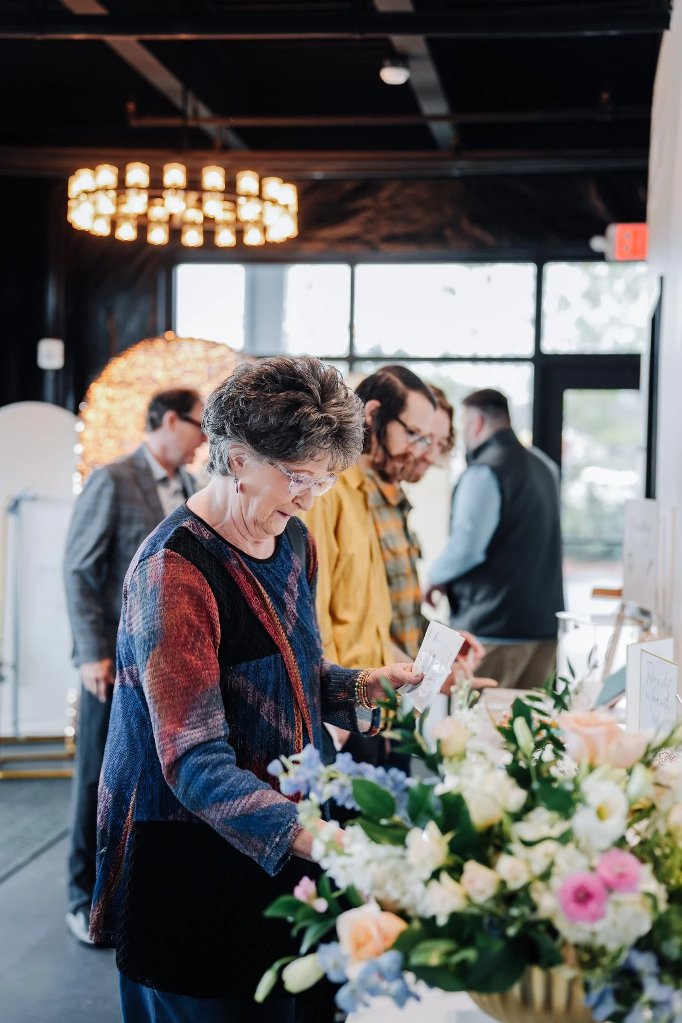 People at a floral display, looking closely at the flowers and literature, in an indoor event space.