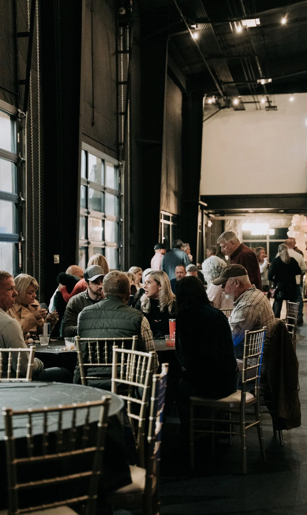 People sitting and chatting at a round table in a dimly lit indoor venue with large windows and a high ceiling.