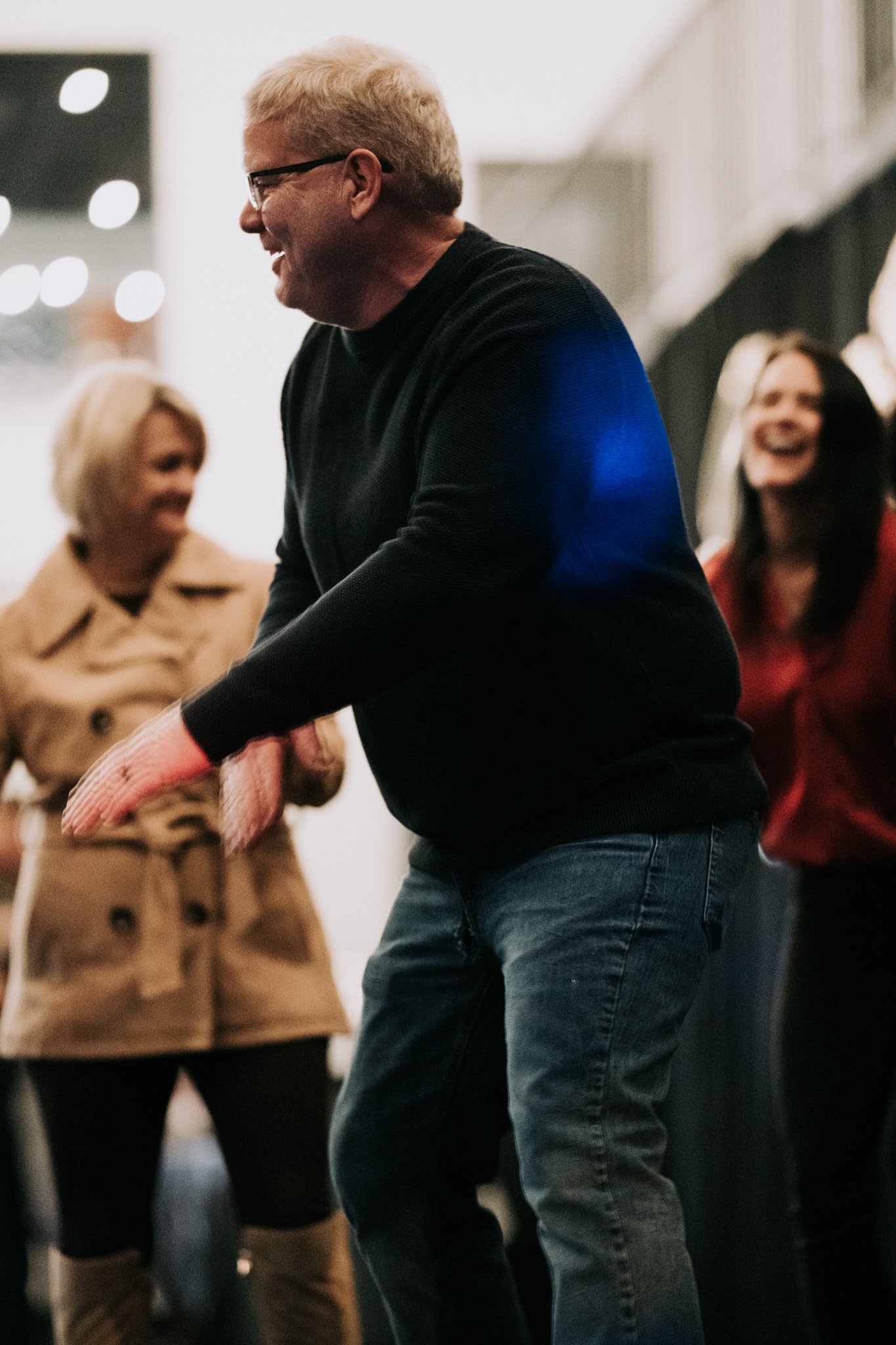 A group of people, including a man wearing glasses, a black sweater, and jeans, dancing and laughing in an indoor setting with blurred background.