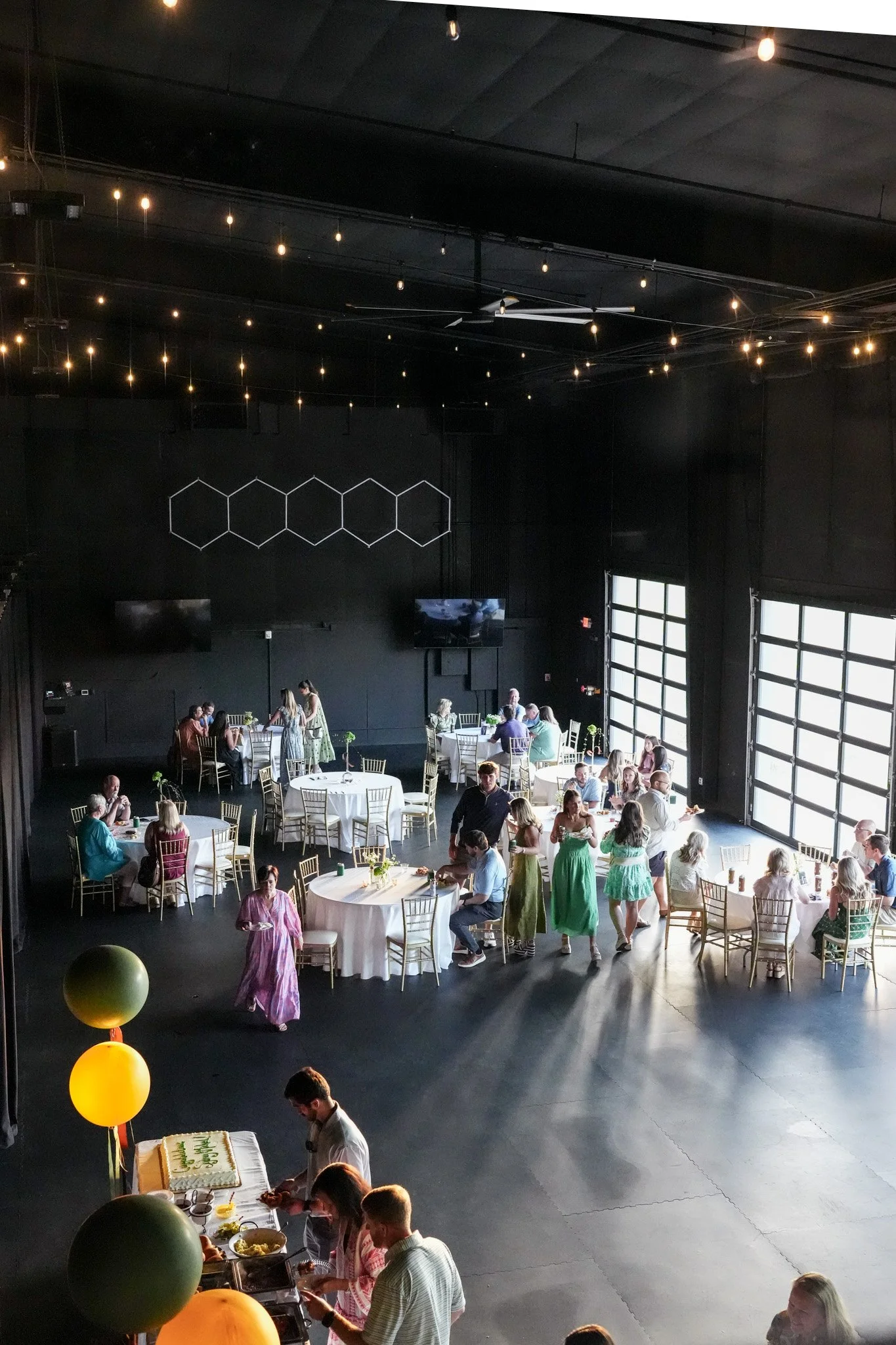 Indoor celebration with guests seated at round tables, some standing and chatting, decorated with balloons and greenery, in a dark hall with string lights and large windows.