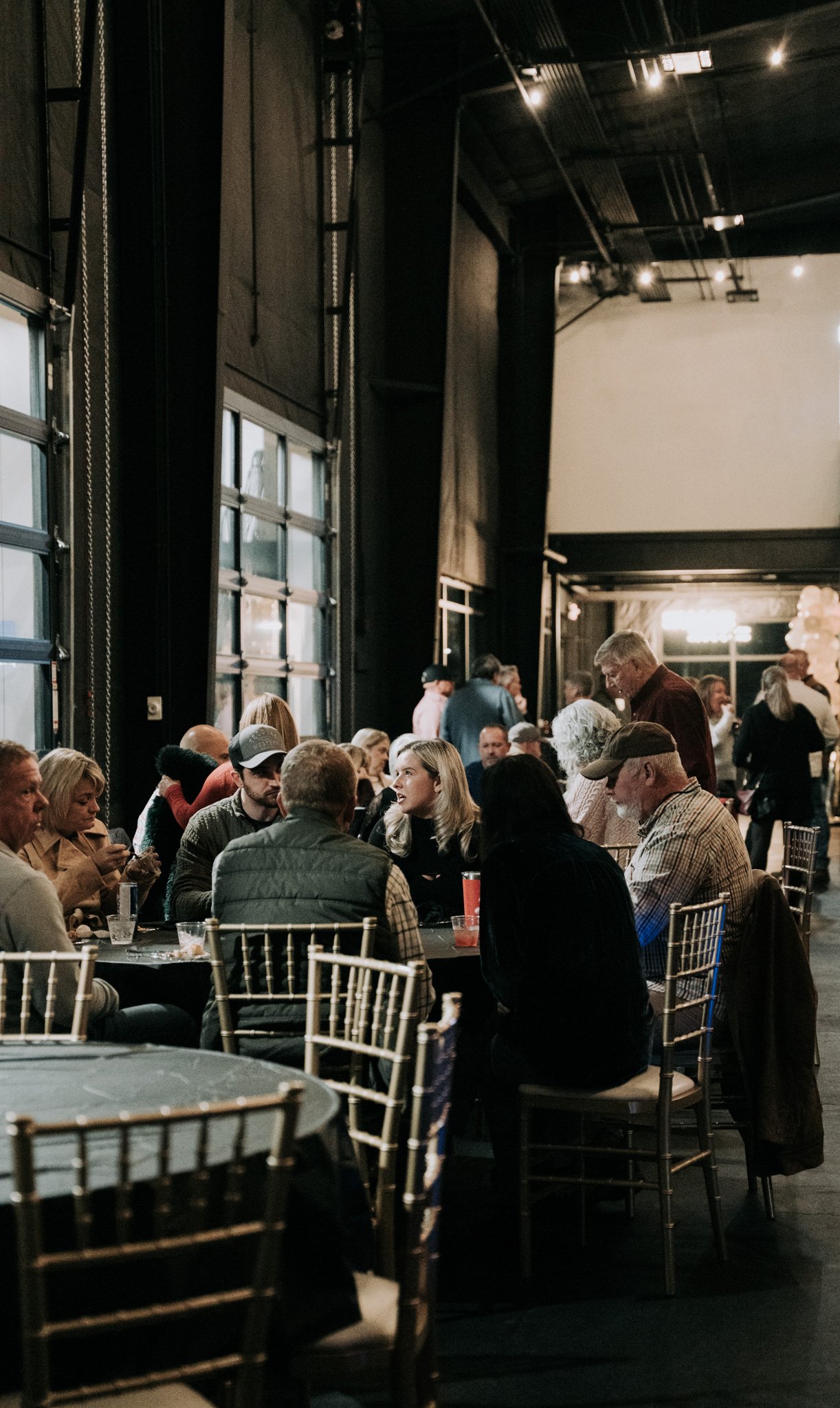 People sitting and socializing at tables in a dimly lit indoor event space with large windows and industrial decor.