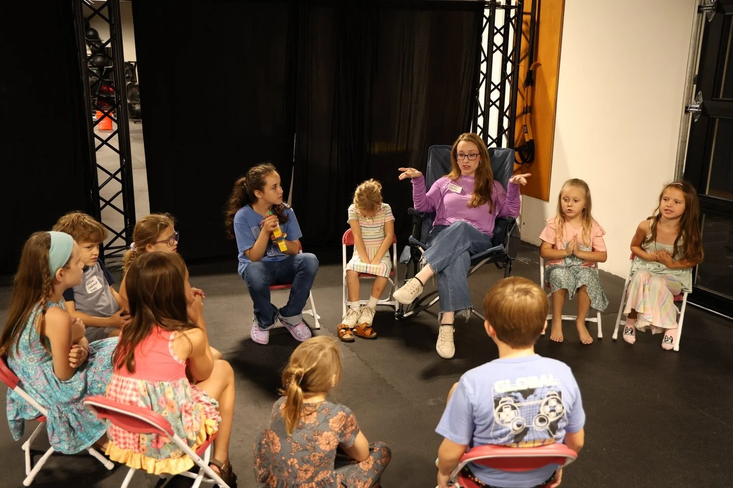 A woman with glasses and long red hair, wearing a purple shirt, sits in a director's chair and appears to be speaking or explaining something to a group of young children seated in a semi-circle around her in a room with black curtains and stage equi