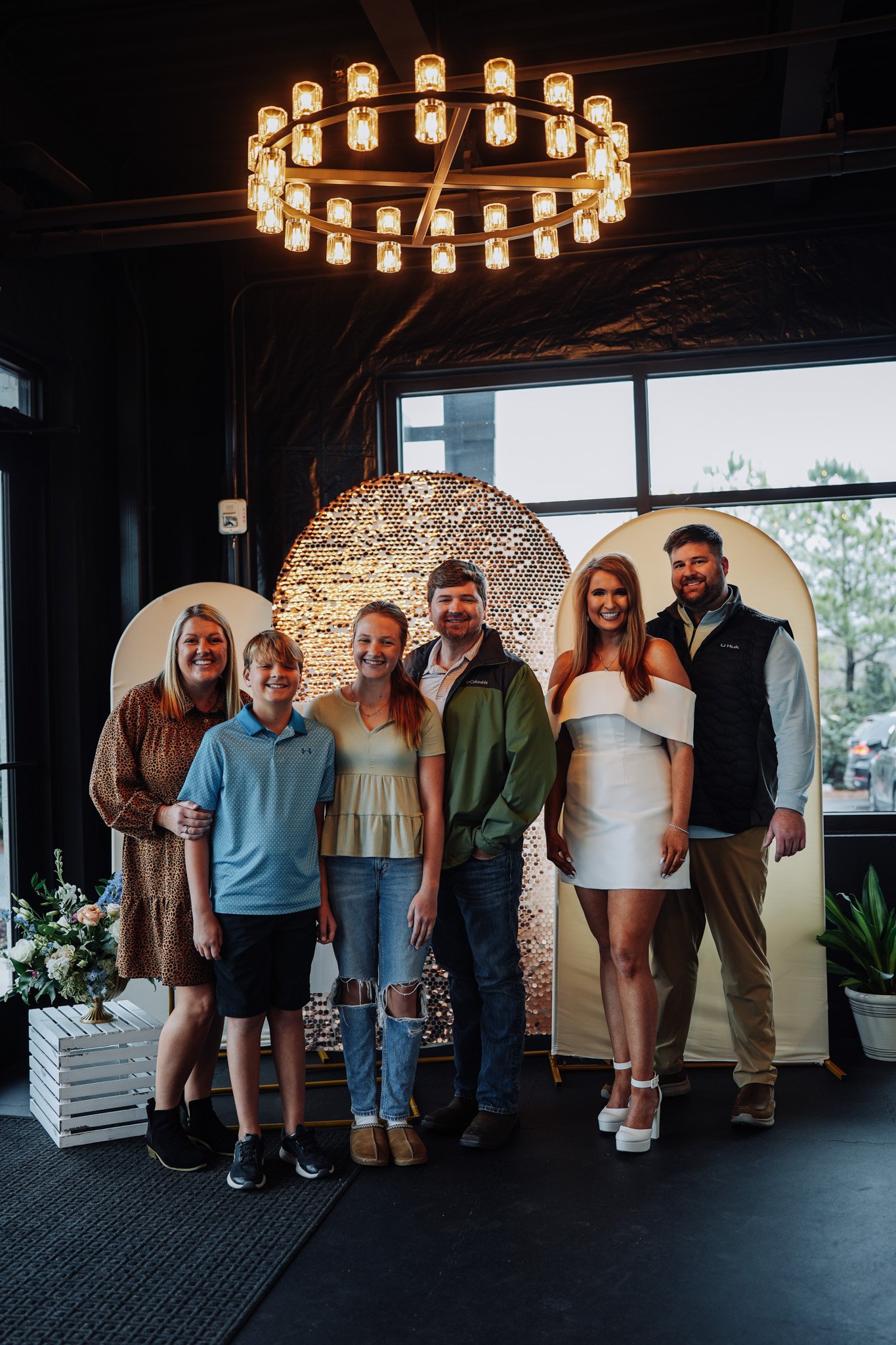 A group of seven people standing indoors in front of a decorative backdrop, including a large circular art piece and floral arrangements, with a chandelier hanging above.