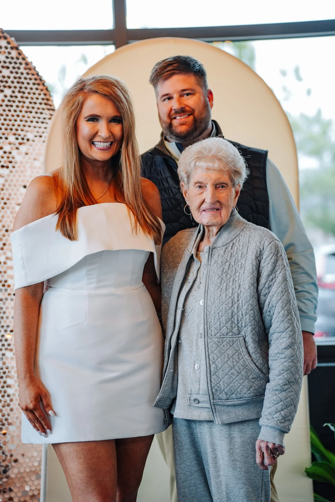 Four people, three women and one man, are smiling and standing together inside a building with glass windows, posing for a photo. The youngest woman is wearing a white dress, the elderly woman is wearing a gray jacket, the man is behind the elderly w