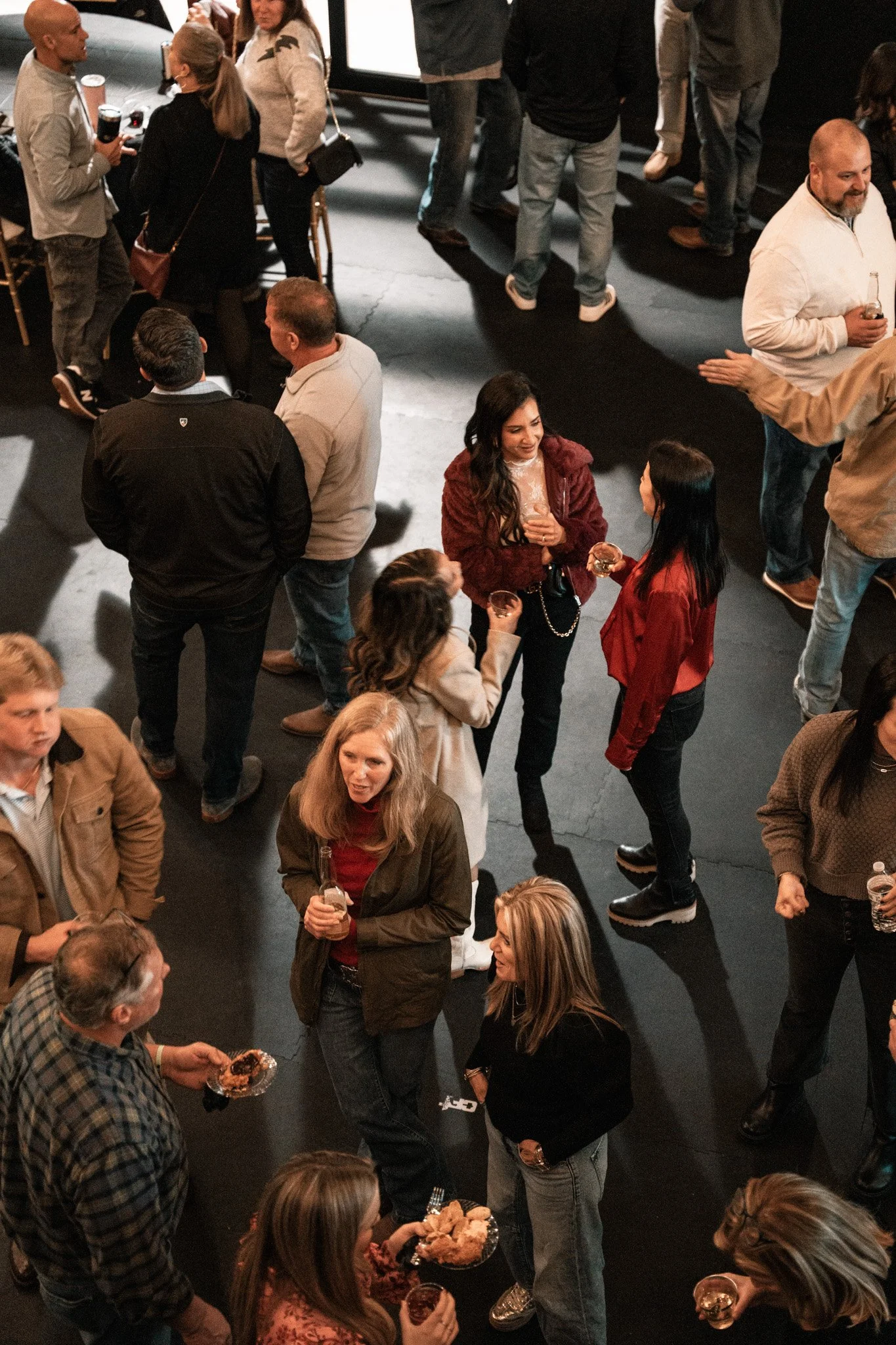 People socializing at a party, some holding drinks and plates of food, on a dark indoor floor with a few standing and chatting in small groups.