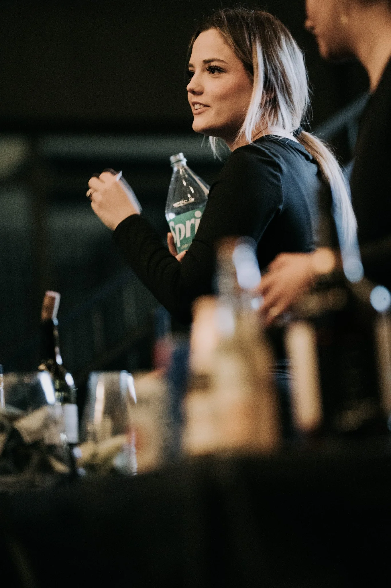 A woman holding a Sprite bottle and a notebook, standing at a bar counter with various bottles and glasses in the foreground.