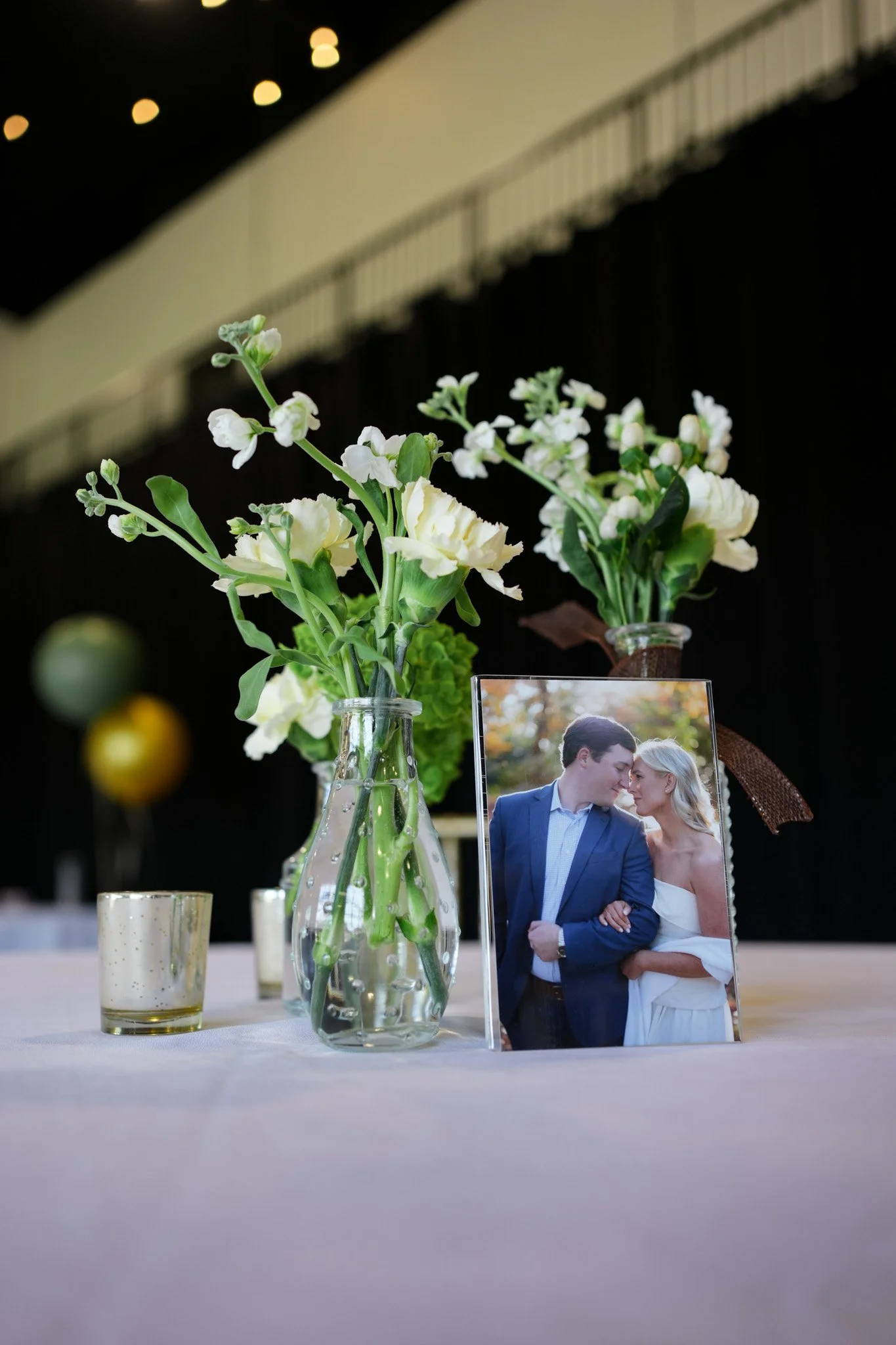 A table decorated with white flowers in a clear vase, a wedding photo of a couple, and some candles, all set in a dimly lit event space.