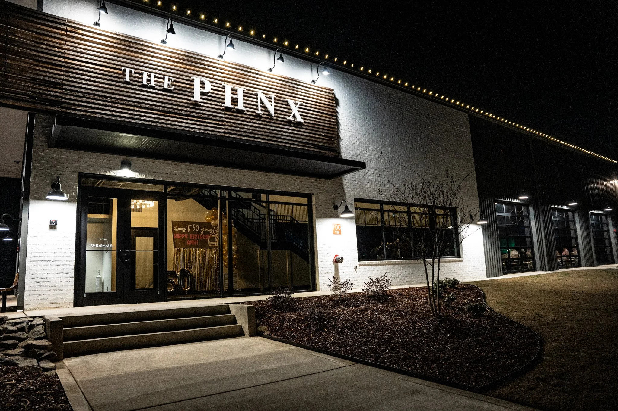 Night view of the exterior of The Pinx restaurant, with its name illuminated on a wooden sign, large glass doors, outdoor lighting, and windows showing indoor seating.
