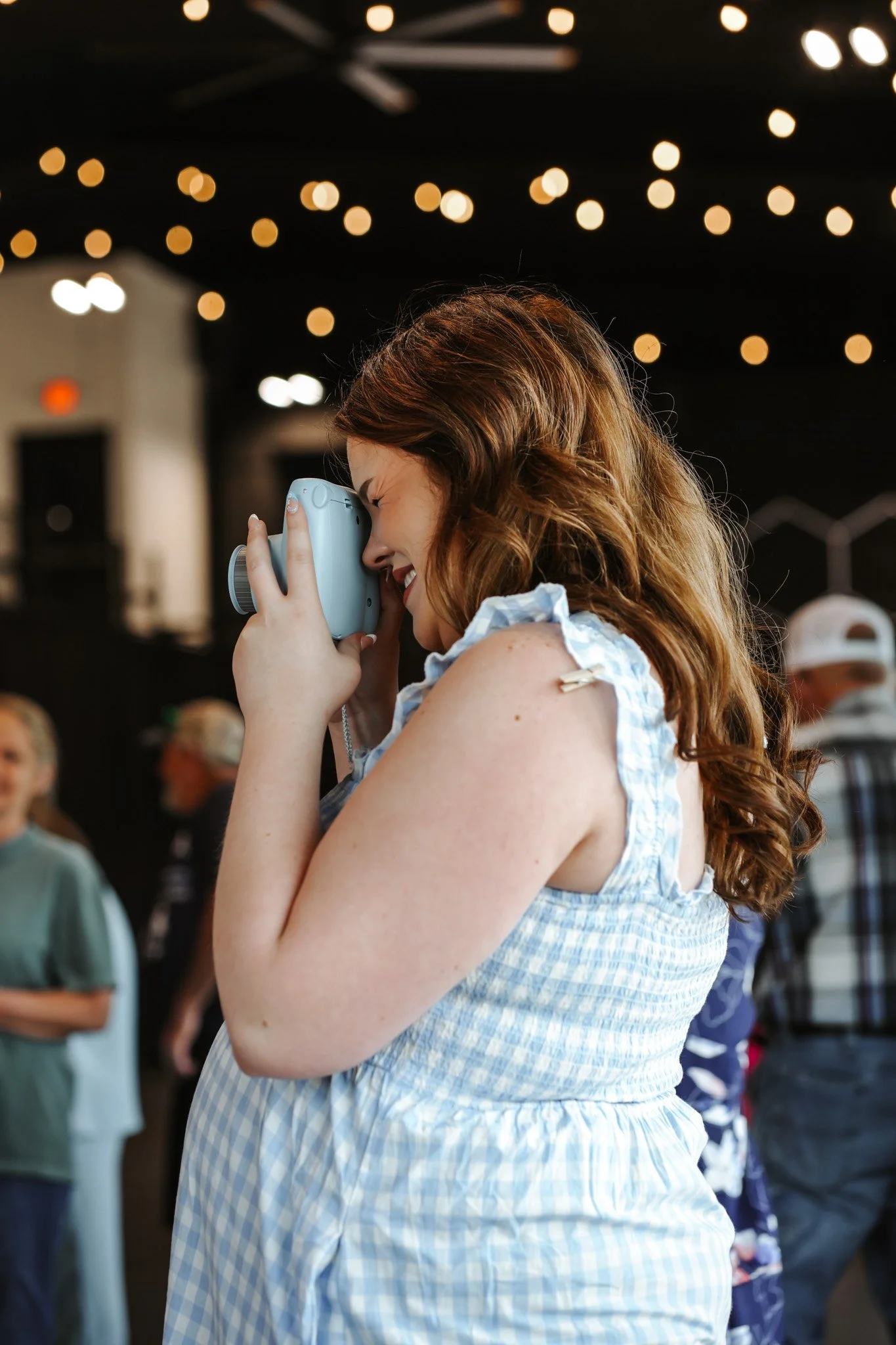 A woman with long, wavy red hair is smiling and holding a vintage camera close to her face, likely taking a picture. She is wearing a blue and white checkered sleeveless dress, and the background shows multiple blurred people and string lights, sugge