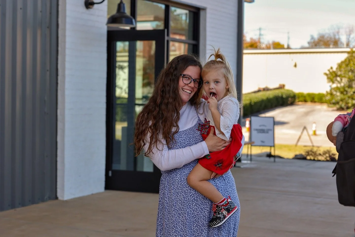 A woman with long curly hair and glasses holding a young girl dressed in a holiday outfit outside a building.