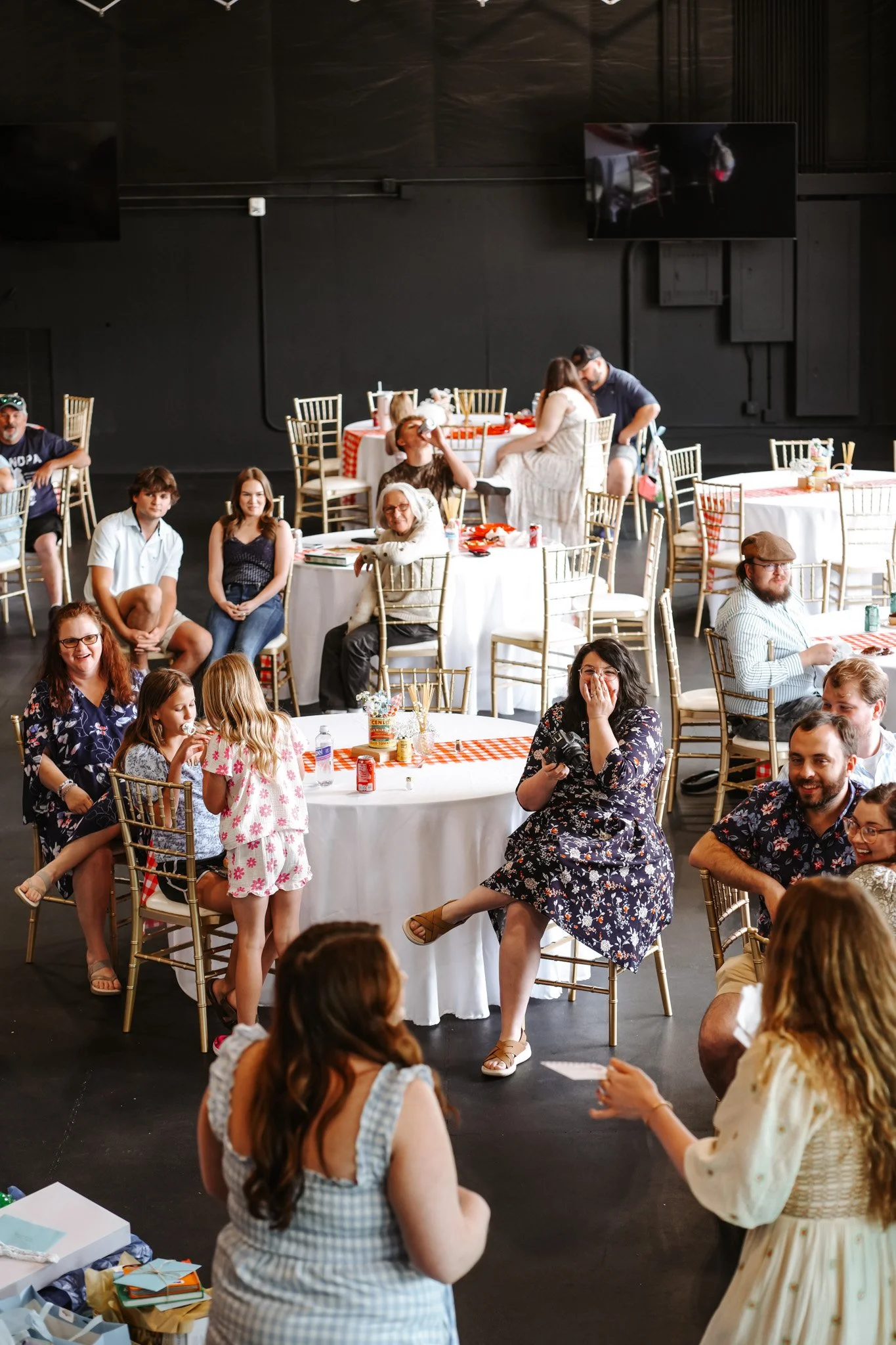 A large indoor gathering with many people engaging in conversations. Some individuals are sitting, while others are standing, in a decorated space with round tables, chairs, and a few displayed items on a table in the foreground.