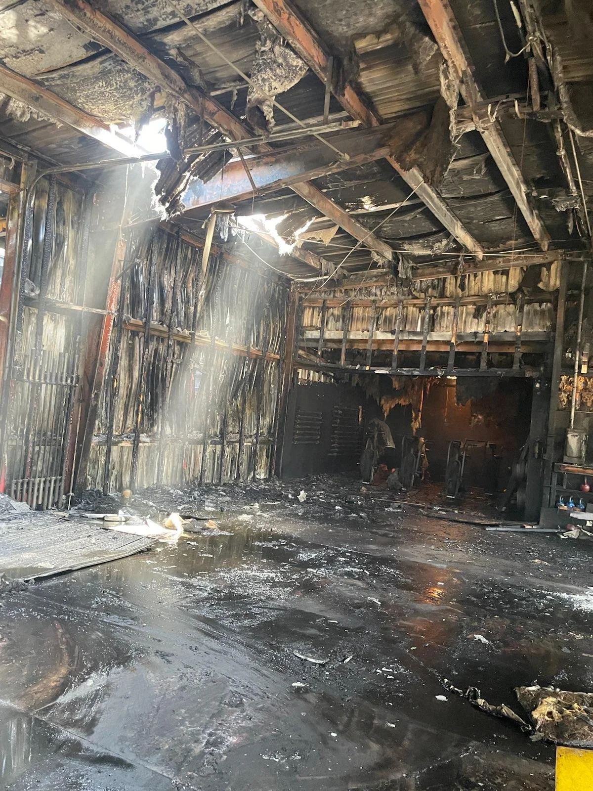 Interior of a house with severe fire damage, charred walls, burnt ceiling, and debris scattered on the wet floor.
