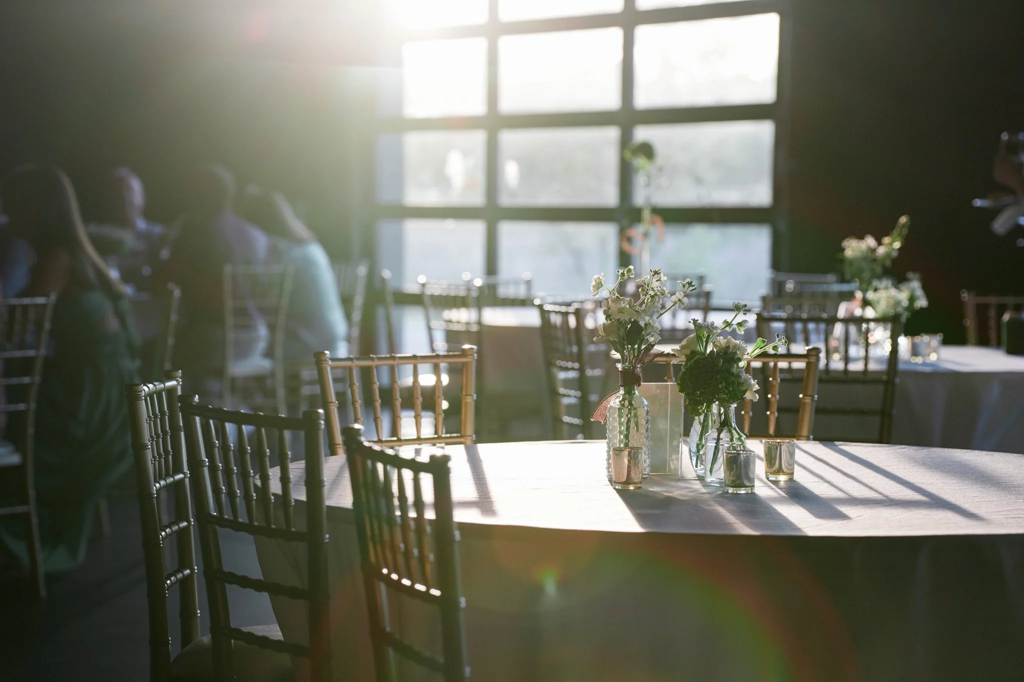 Sunlight streaming through a large window illuminating a dining area with several wooden chairs and a table decorated with small vases of flowers.