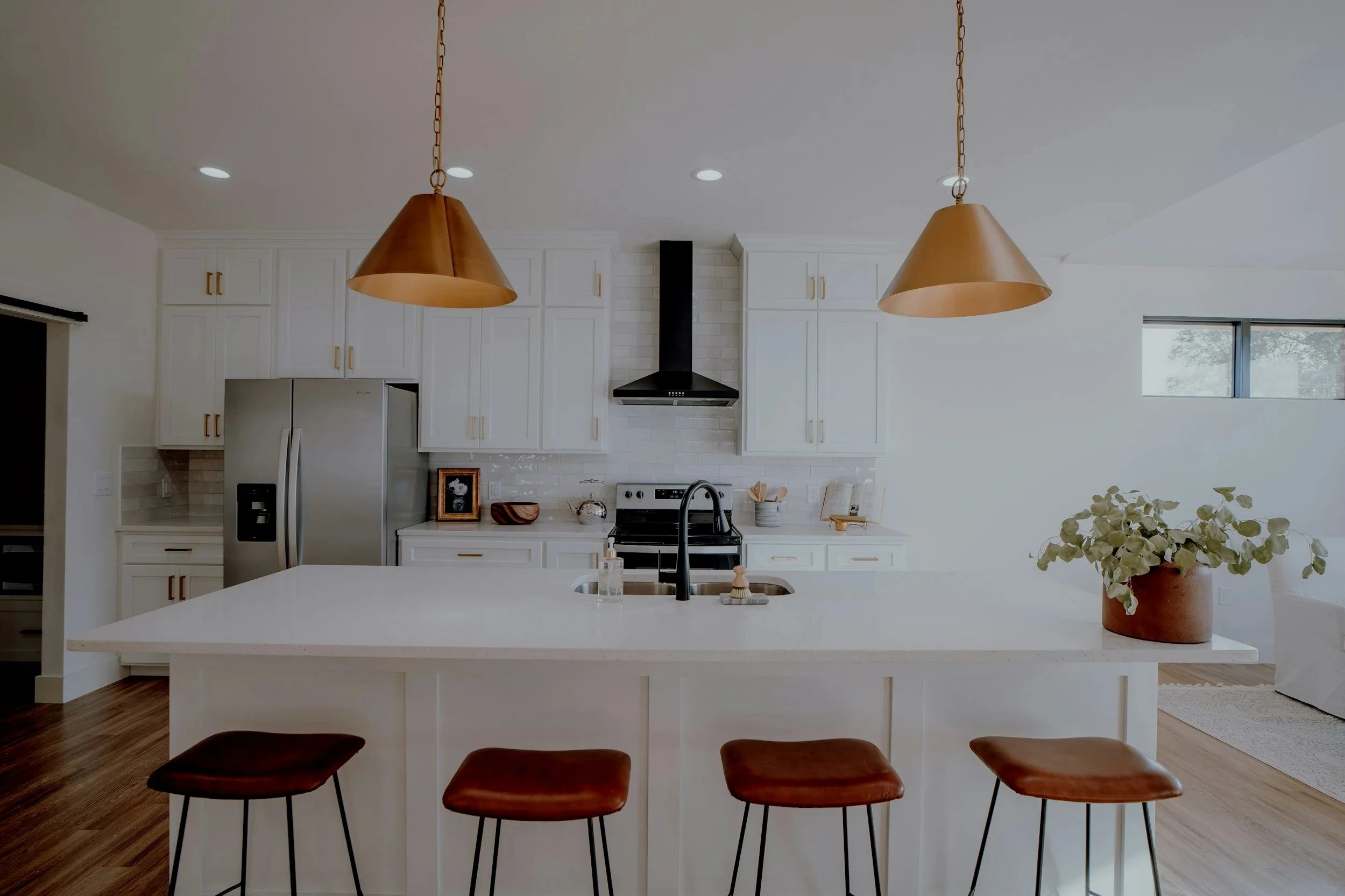 Modern kitchen with white cabinetry, a stainless steel refrigerator, black stove with a black vent hood, a white countertop island with four brown leather bar stools, pendant lights, and a potted plant.