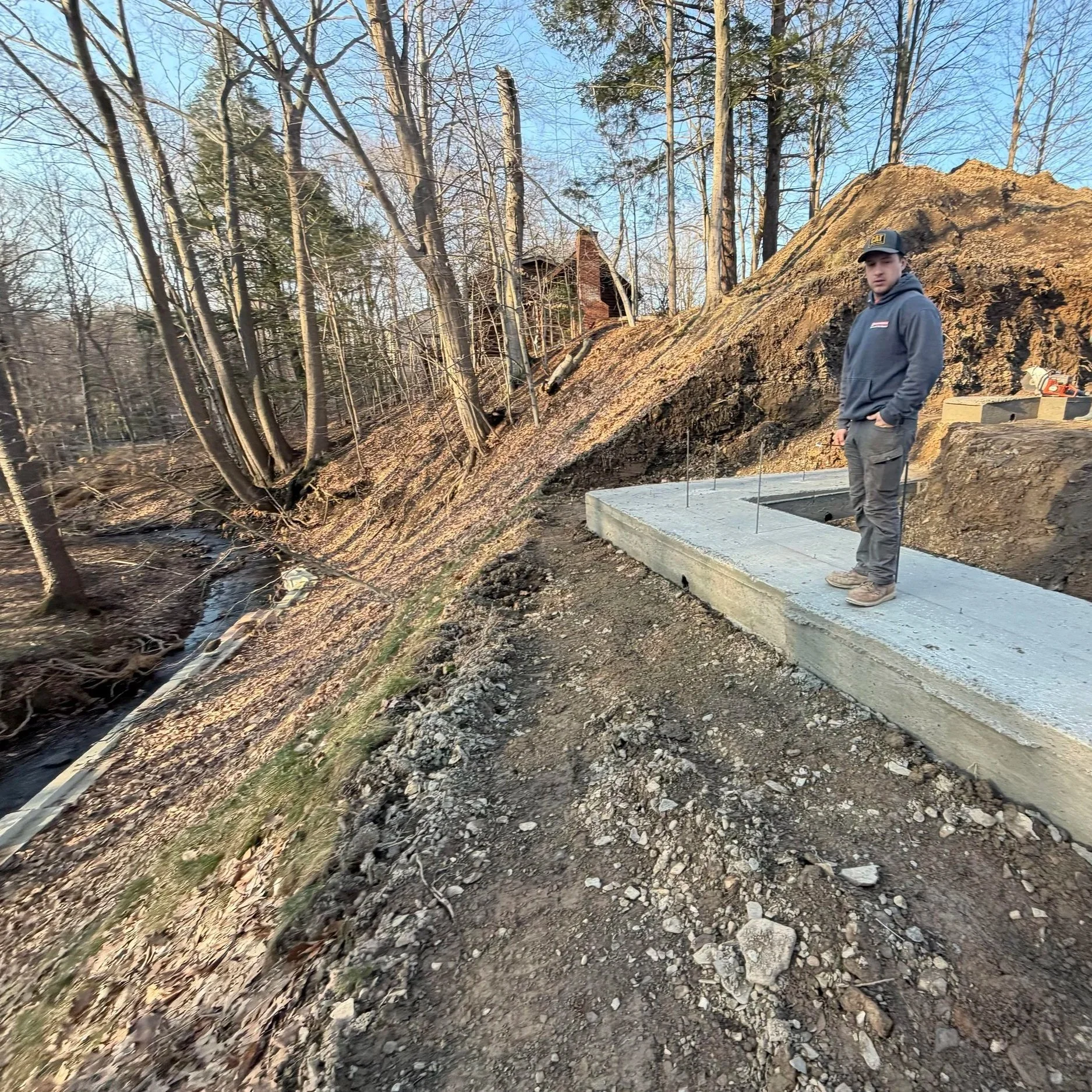 A construction worker stands on a freshly poured concrete slab on a hillside, with exposed soil and a small stream running alongside. A wooded area with barren trees and a cabin on a hill are visible in the background during daytime.