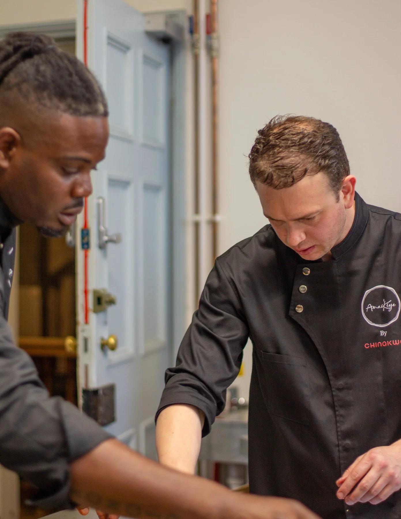 Two chefs working together in a kitchen, wearing black chef jackets. One chef is focused on preparing food, with a serious expression, while the other is assisting or observing.