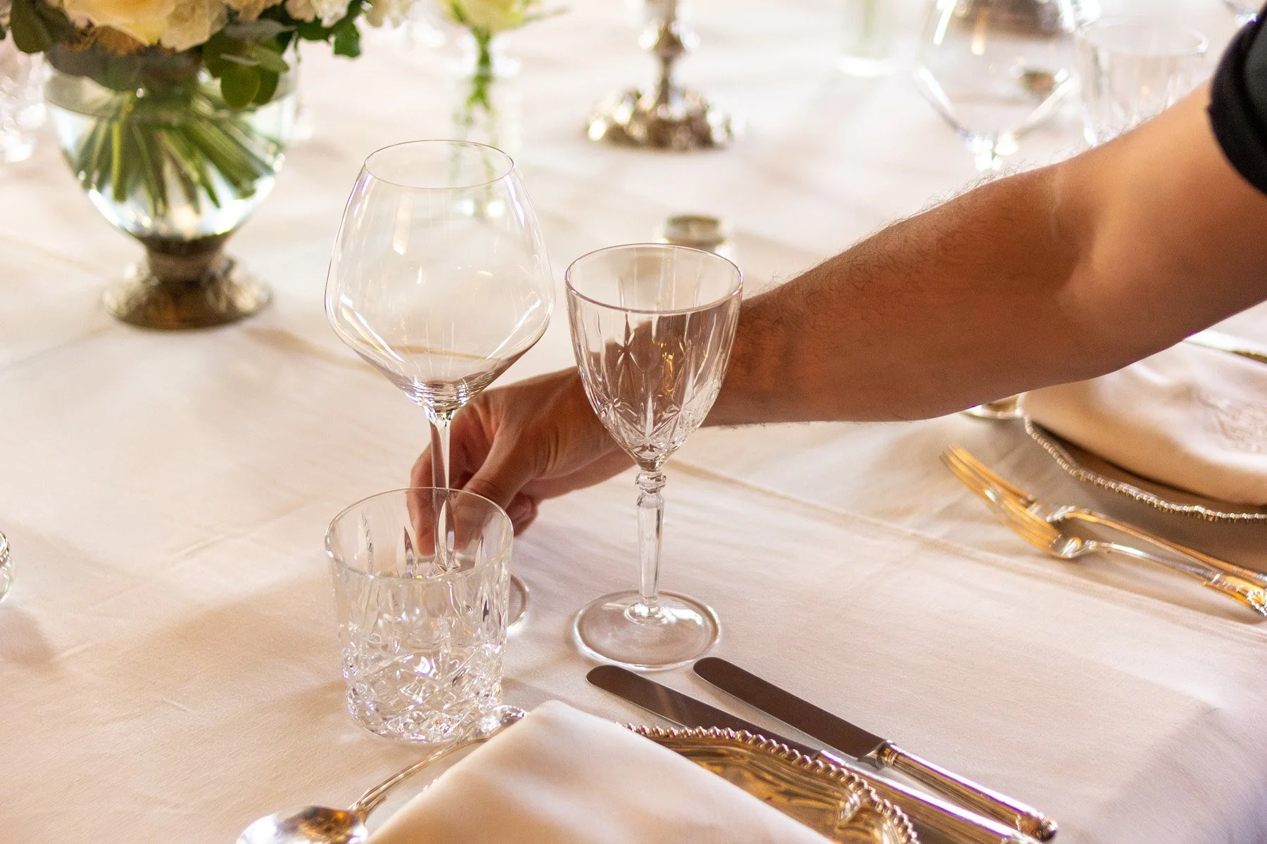 A person's hand placing a wine glass on a formal dining table with crystal glassware, silverware, plates, and a floral centerpiece.
