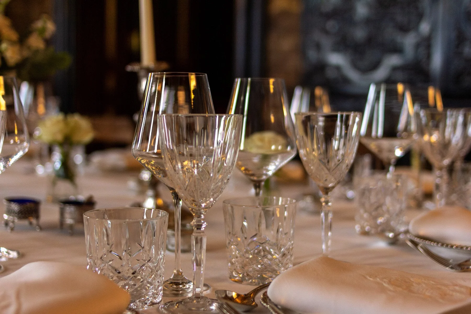 Elegant table setting with multiple glassware including wine glasses and tumblers, beige napkins, and cutlery, likely prepared for a formal dinner event.