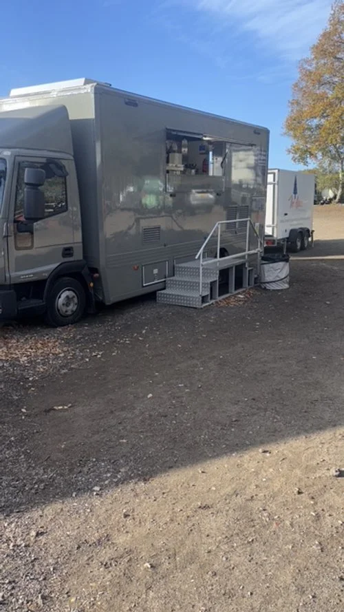 Gray food truck with a small set of stairs leading to serving window, parked on dirt ground with another trailer in the background and a tree with fall foliage.