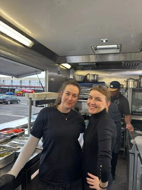 Two women standing inside a commercial kitchen, smiling at the camera, with a man cooking in the background.