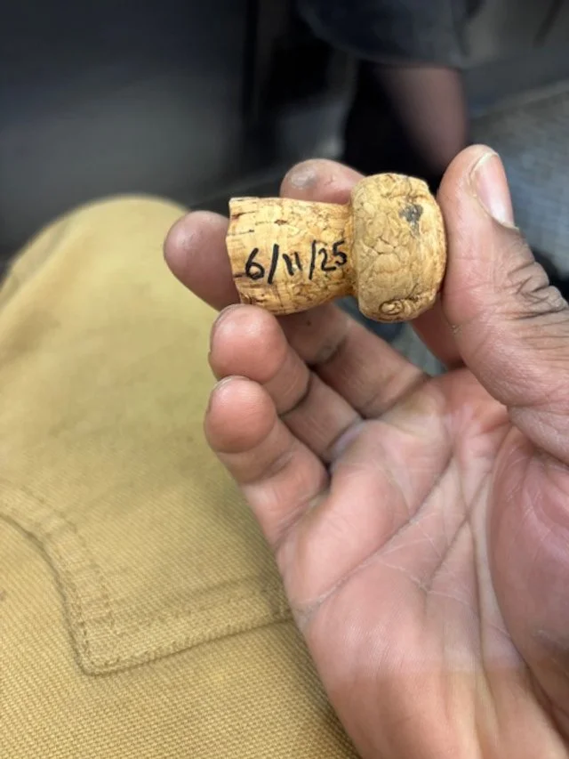 Close-up of a person's hand holding a small, carved cork with the handwritten date '6/1/25' on it, against a background of tan fabric and a blurred person's arm.