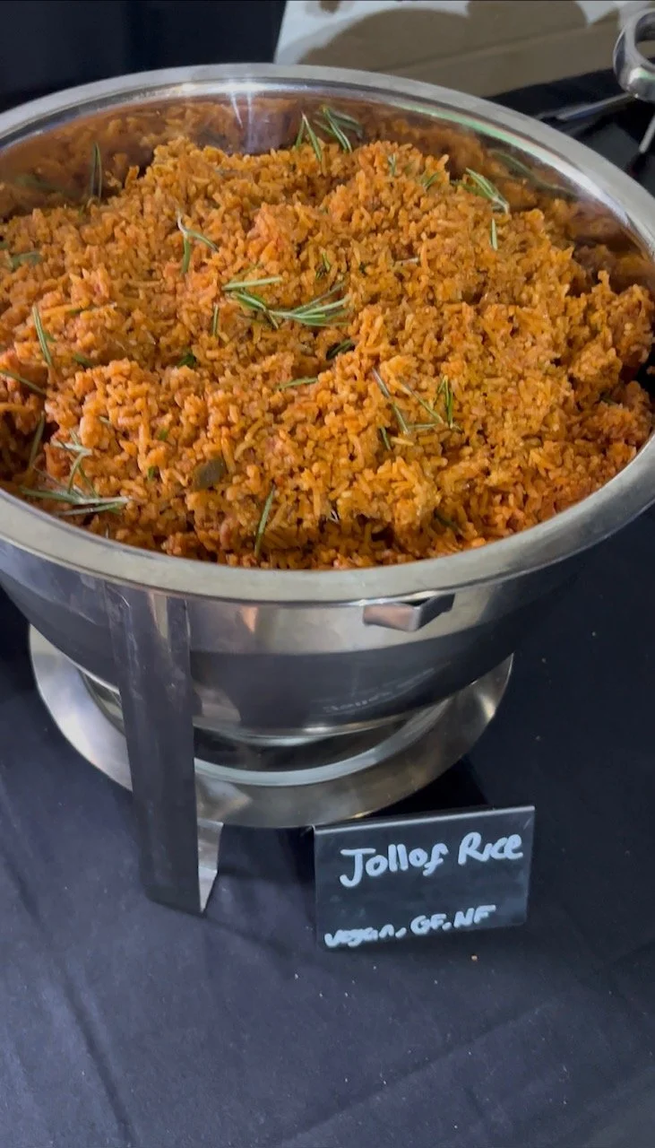 A large stainless steel serving dish filled with jollof rice garnished with fresh herbs, on a black table with a small black sign indicating 'Jollof Rice' and 'Vegan, GF, NF' indicators.