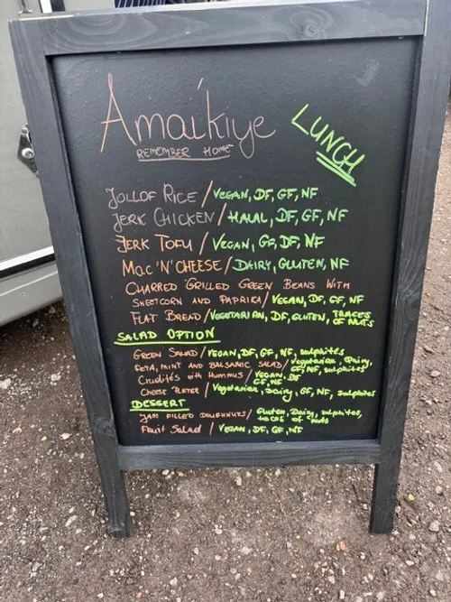 Menu board from Ama Kuiye restaurant listing vegan, gluten-free, and healthy dishes, including jerk chicken, jerk tofu, and mac n' cheese with options for salads and desserts.