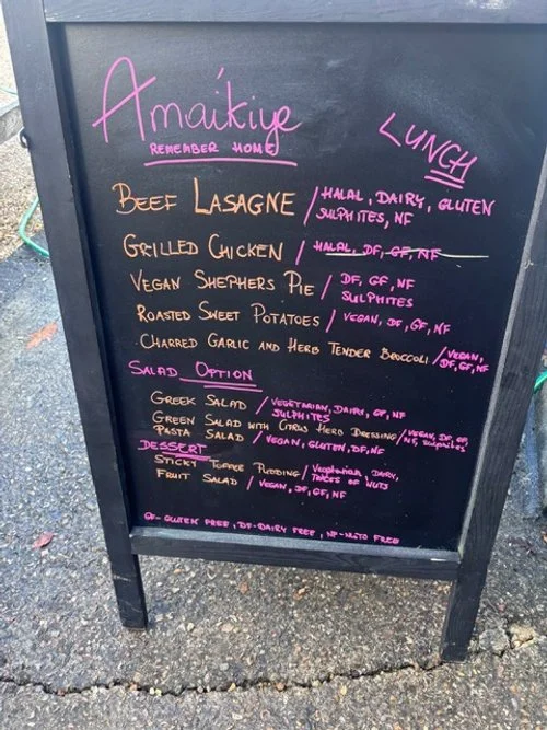 Black chalkboard menu featuring a vegan restaurant's offerings, including beef lasagne, grilled chicken, vegan shepherd's pie, roasted sweet potatoes, charred garlic with broccoli, and various salads and desserts, with dietary notes