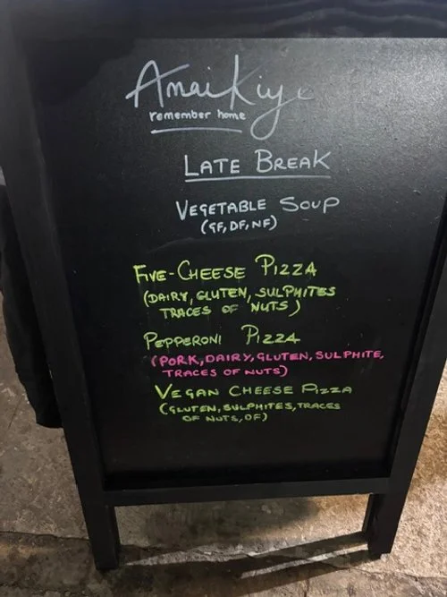 A chalkboard menu outside a restaurant with the title 'Amaze' and the subtitle 'remember home'. It lists items including late break, vegetable soup, five-cheese pizza, pepperoni pizza, and vegan cheese pizza, with allergy notes in parentheses.