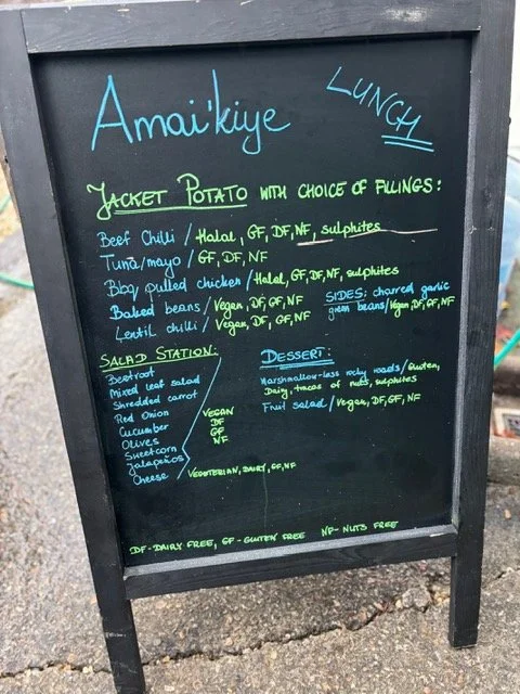 Menu board featuring a jacket potato with various filling options, salad station with vegetables, and dessert choices, written in colorful chalk on a blackboard.