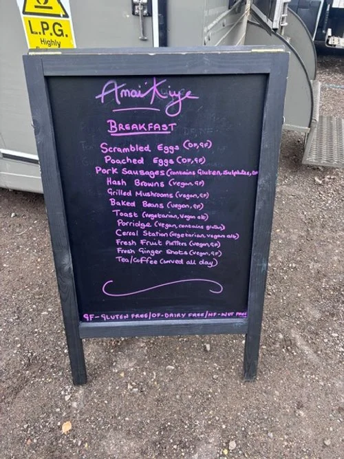 Blackboard menu listing breakfast items including scrambled eggs, poached eggs, pork sausages, hash browns, grilled mushrooms, baked beans, toast, porridge, cereal, fresh fruit, fresh ginger shots, and tea or coffee. Handwritten in purple and pink ch