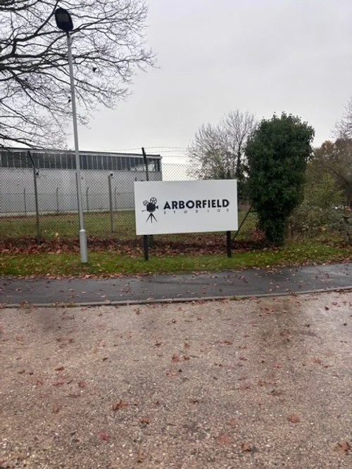 Sign that reads 'Arborfield Studios' with a film reel and camera icon, outside in a parking lot area with a chain-link fence and trees in the background.