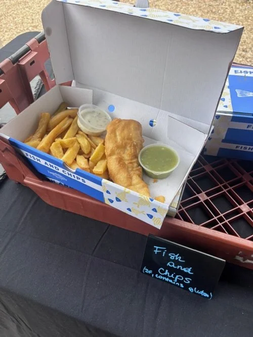 A cardboard box containing fish and chips with fries, tartar sauce, and tart green dipping sauce, placed on a black table with a chalkboard sign.