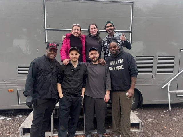 Group of seven people standing outdoors in front of a large gray vehicle, some smiling and posing for the camera, with three people in the front row and four behind.