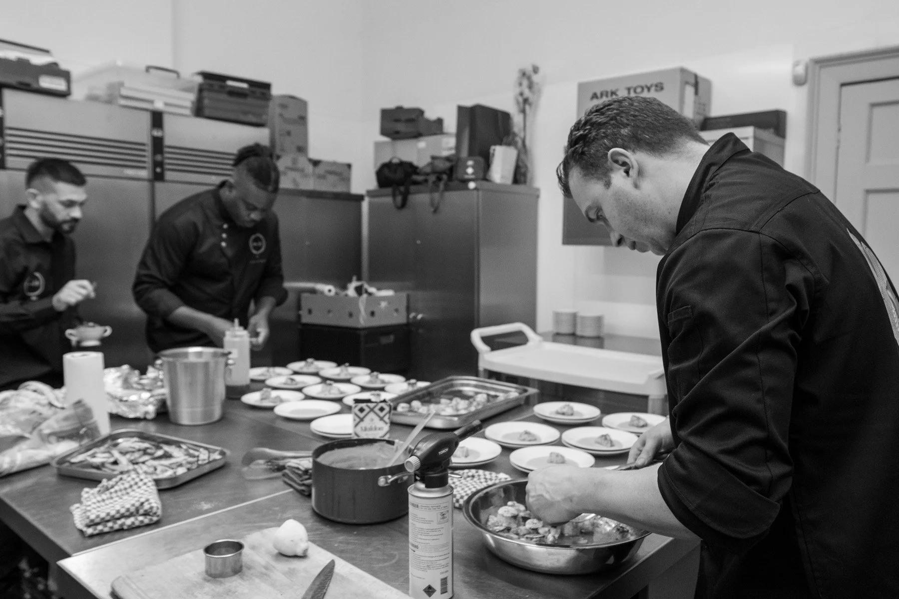 Three chefs are preparing food in a kitchen, with plates laid out on the counter and various cooking tools and ingredients visible.