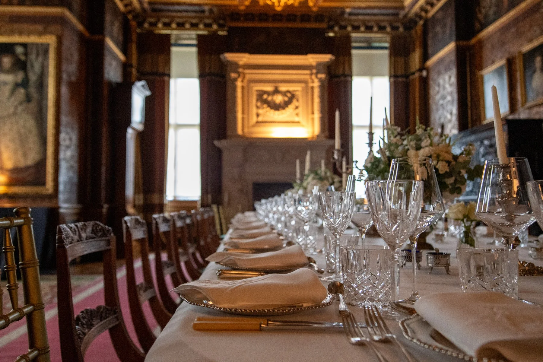 Elegant dining room with a long table set with white napkins, crystal glassware, and floral centerpieces, in a room with dark wood paneling, paintings, and large windows.