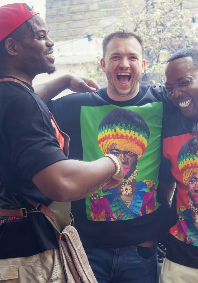 Three men are smiling and laughing, with their arms around each other, wearing matching T-shirts featuring a woman with a colorful headwrap and jewelry, outdoors with a brick wall and trees in the background.