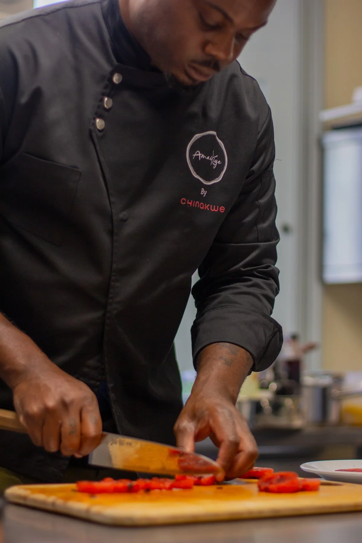 A person in a black chef's coat is slicing red bell peppers on a wooden cutting board in a kitchen.