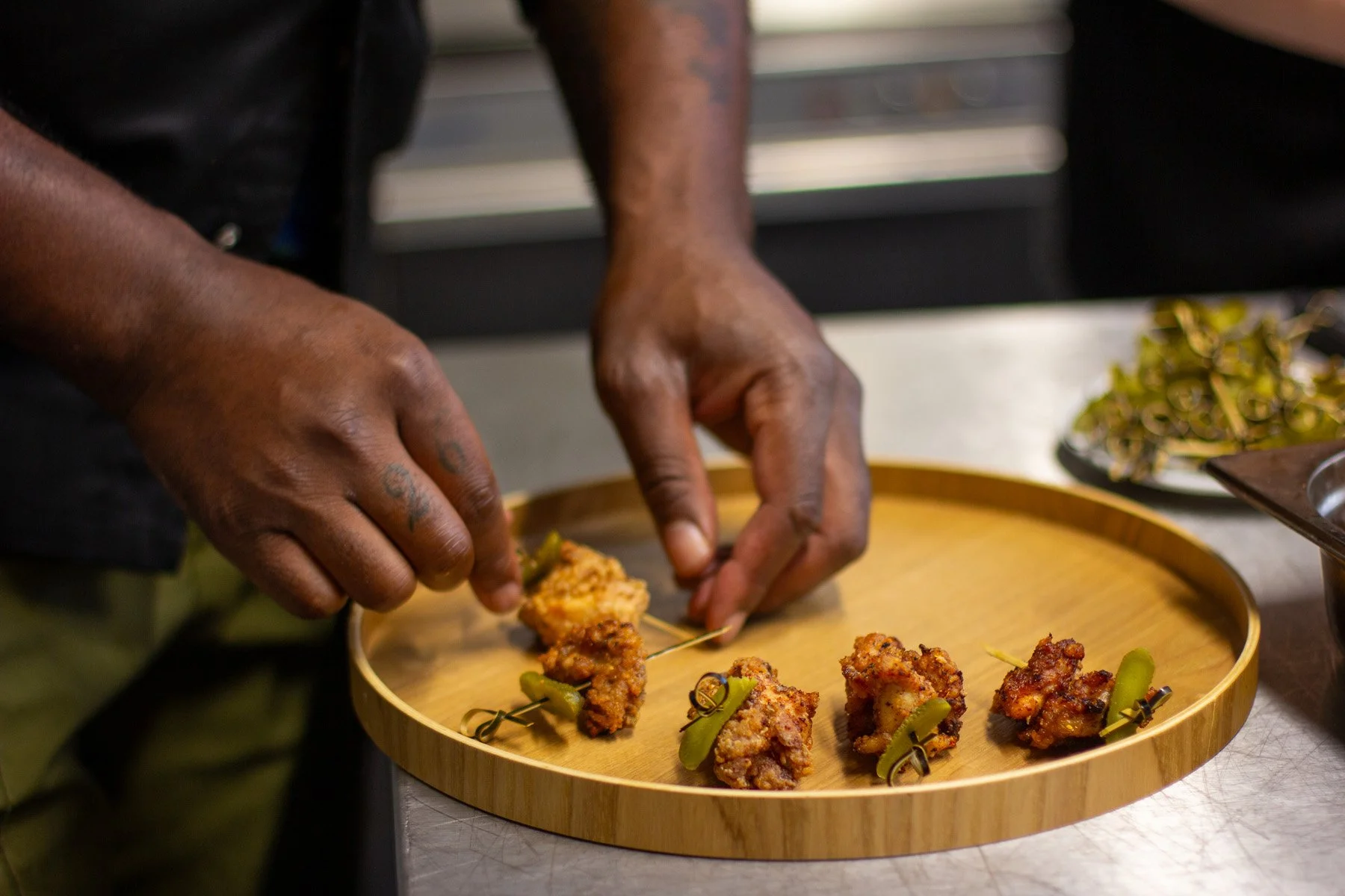 A person arranges pieces of fried chicken with green chili peppers on a round wooden serving tray.