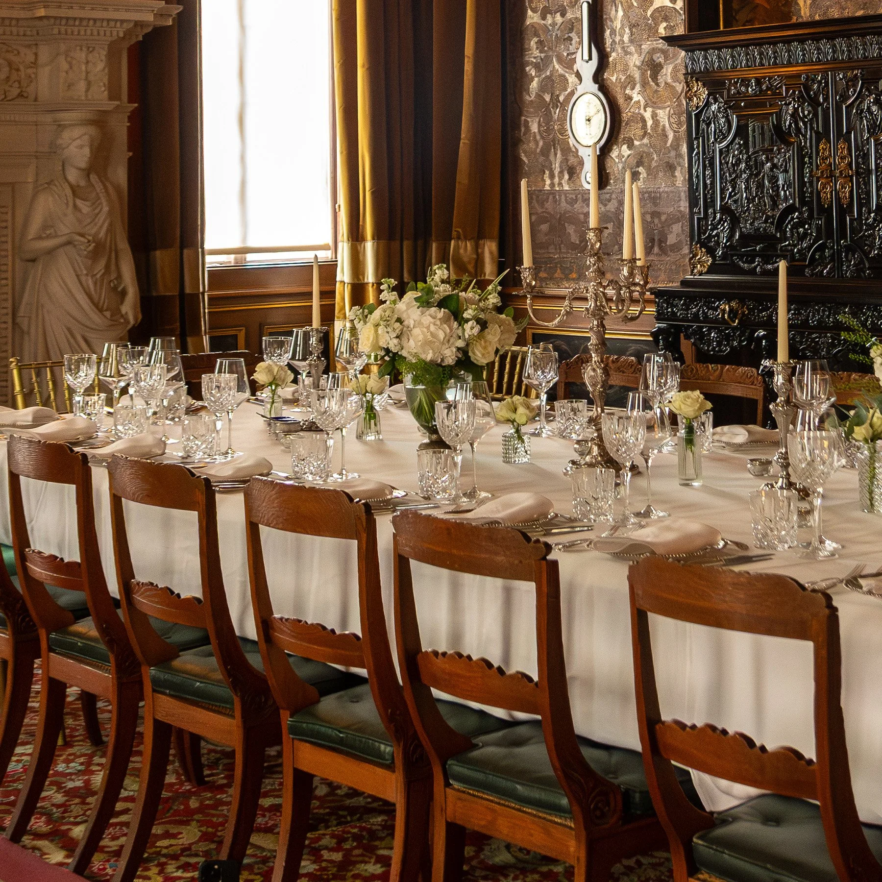 An elegant dining room with a long table set with white linens, crystal glasses, silverware, and white flower centerpieces. The room features dark wooden furniture, gold curtains, a candelabra, and a large ornate cabinet, with a window letting in nat