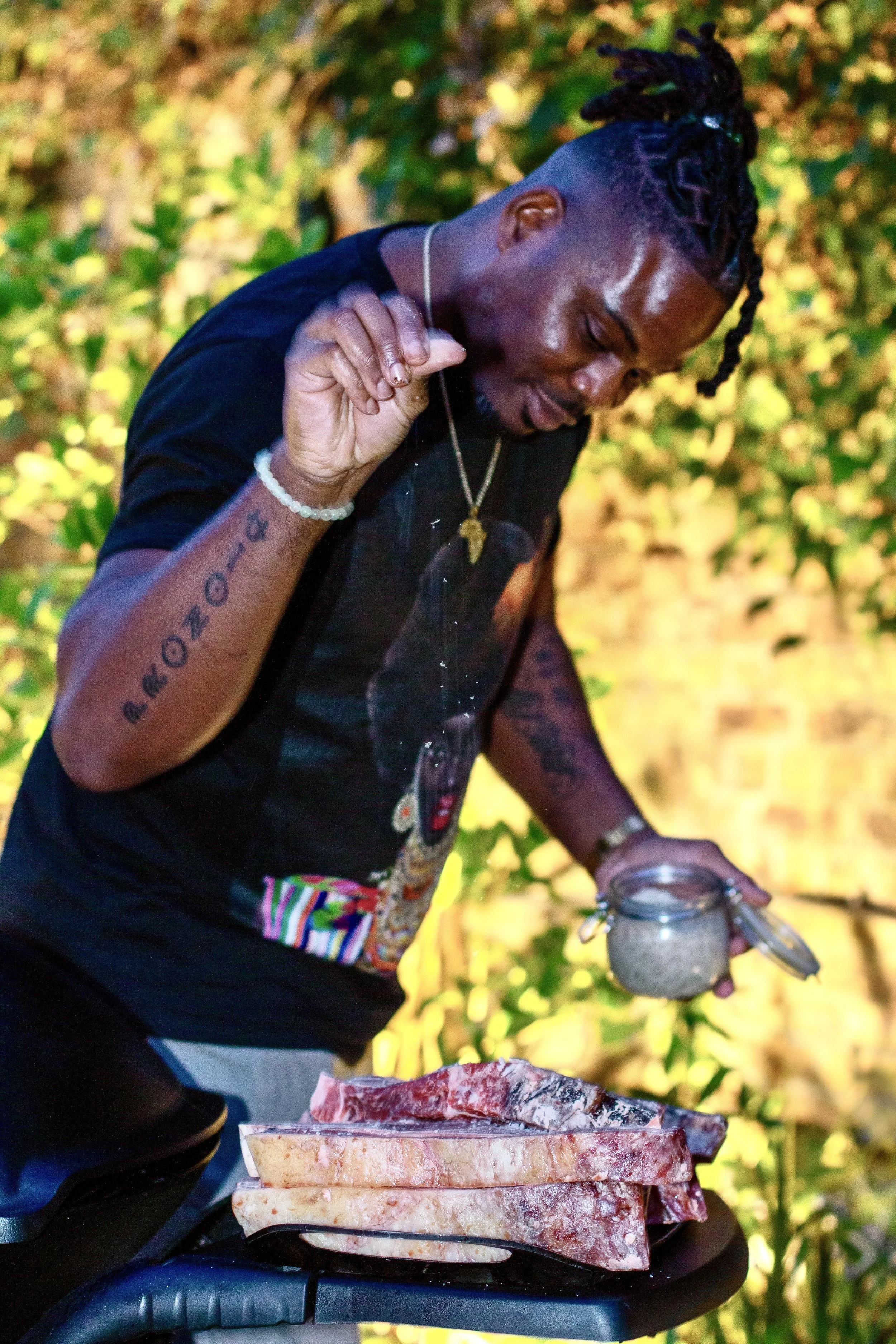 A man with dreadlocks wearing a black T-shirt and jewelry, standing outdoors with green foliage in the background, holding a jar of seasoning and seasoning a large piece of raw meat on a table.
